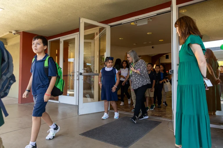 A group of elementary school children in uniforms exit a building through open glass doors, accompanied by two female adults, one holding the door and another standing nearby in a green dress.