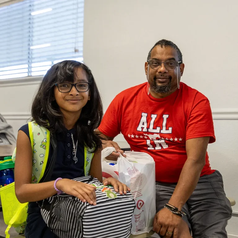A young girl with glasses sits next to an adult man in a red “ALL IN” shirt. They are indoors, smiling, with bags and backpacks around them, suggesting they are packed for travel or an outing.