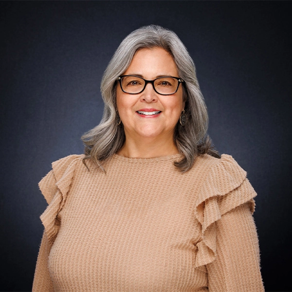 Smiling woman with gray hair and glasses, wearing a textured beige top with ruffled sleeves, standing in front of a dark, plain background.