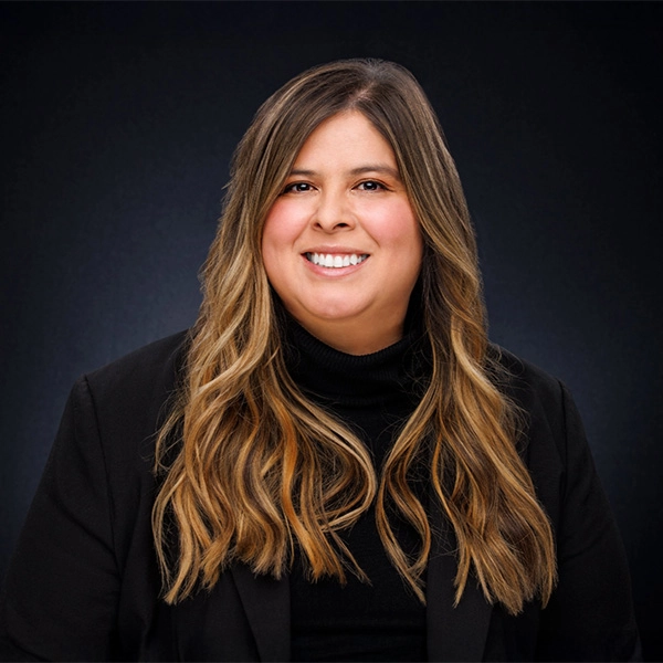 A woman with long, wavy light brown hair smiles at the camera. She is wearing a black blazer and black turtleneck against a dark, gradient background.