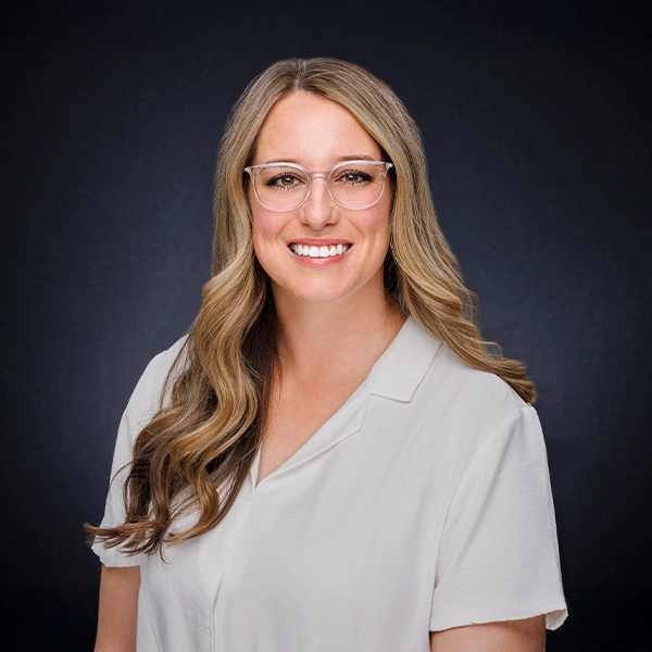 A woman with long wavy blonde hair, wearing clear glasses and a white blouse, smiles at the camera against a plain dark background.