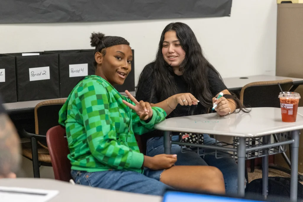 Two students sit at a classroom desk. One, wearing a green hoodie, smiles and flashes a peace sign, while the other, in a black shirt, smiles and holds a marker. A drink and organized folders are on the desk nearby.