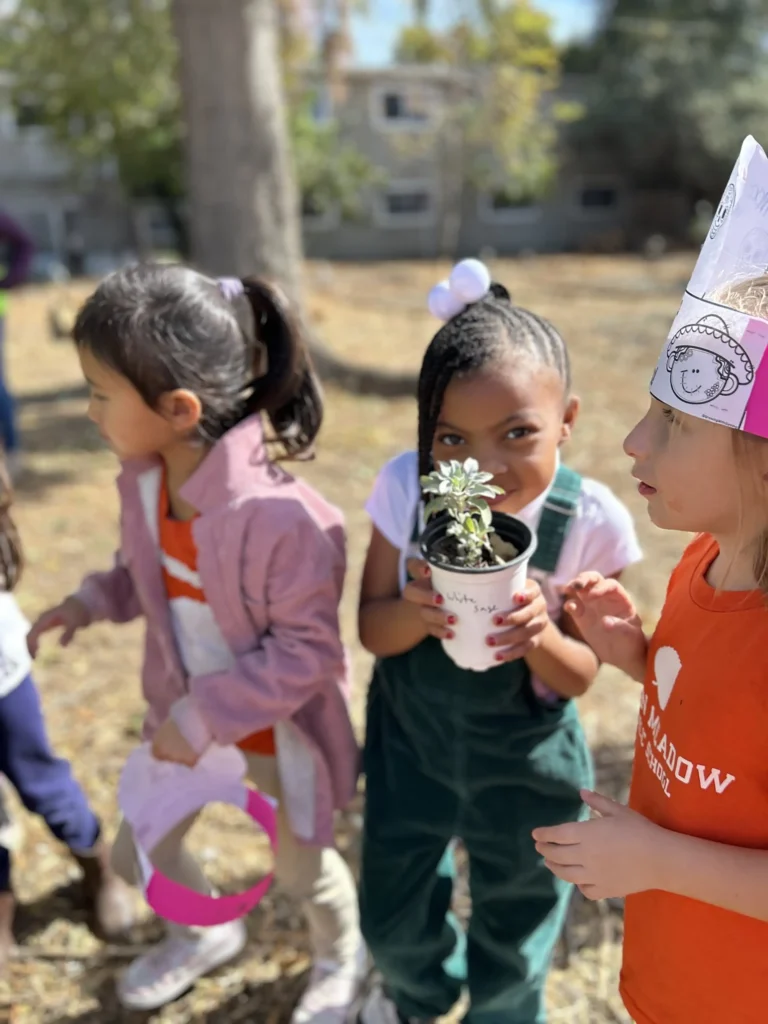 Three young children stand outdoors; one girl holds a potted plant, another wears a paper crown, and the third looks to the side. They appear to be at a school activity or event on a sunny day.