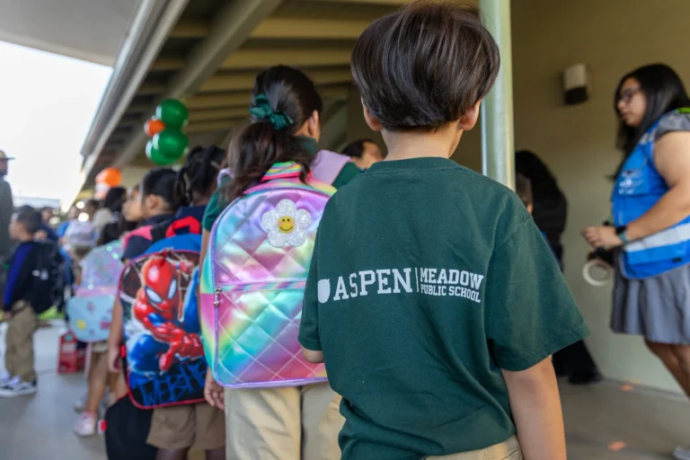 Children wearing backpacks line up outside a school building. One child in the foreground wears a green shirt with Aspen Meadow Public School written on the back. An adult stands nearby, wearing a blue vest.