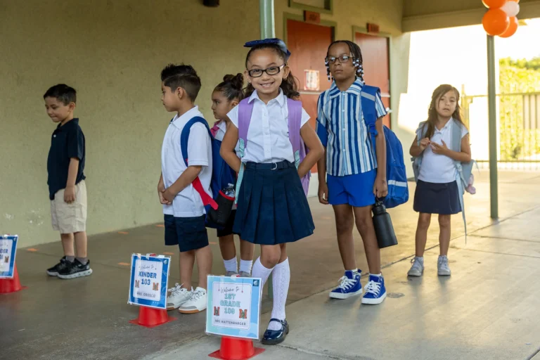 A group of young children wearing school uniforms stands in a line outside a school building, smiling and carrying backpacks. Signs in front of them indicate grade levels.