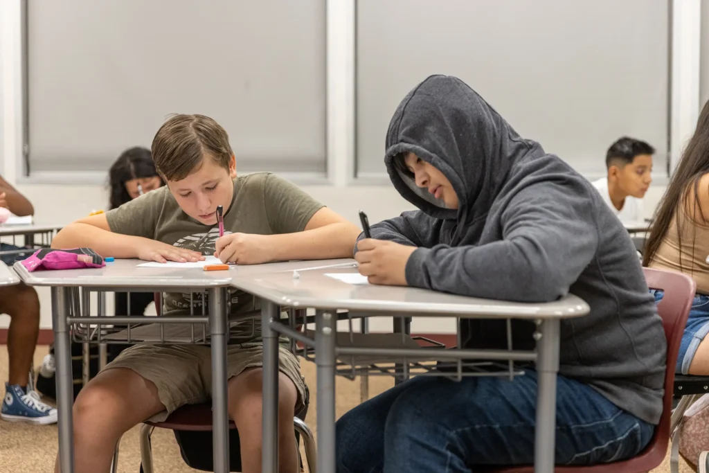 Two students sit at desks in a classroom, writing on paper. One wears a t-shirt and shorts, while the other wears a hooded sweatshirt. Other students are seated and working in the background.