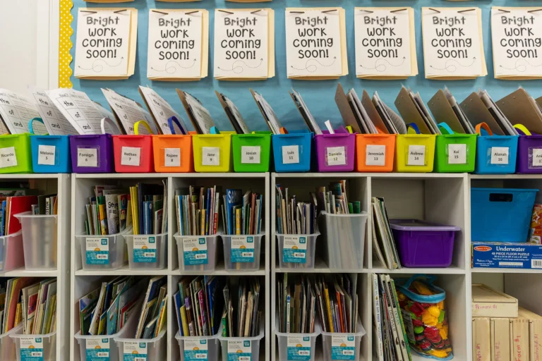 Classroom shelves hold colorful bins of papers and books. Above, signs say “Bright work coming soon!” Bins are labeled for different subjects, and books are organized in labeled cubbies below.
