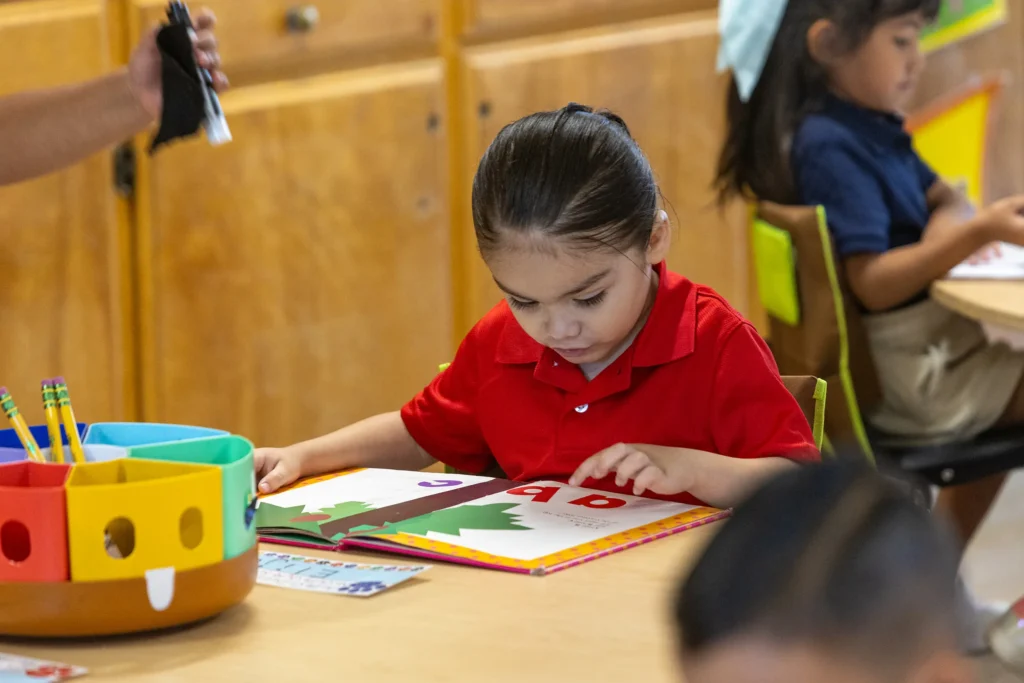 A young child in a red shirt sits at a classroom table, focused on reading a colorful book. Other children are seated at nearby tables, and school supplies are visible on the table in the foreground.
