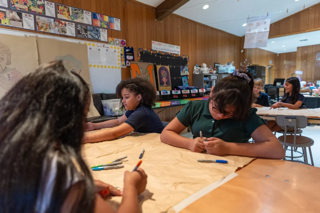 Children sit at tables in a classroom, drawing on large sheets of paper with markers. The room has wooden walls, colorful decorations, and art supplies scattered on the tables.