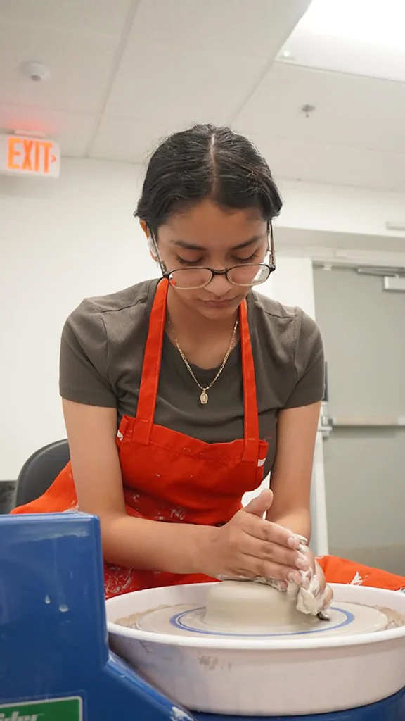 A young woman wearing glasses and a red apron shapes clay on a pottery wheel in a bright room, focused on her work.
