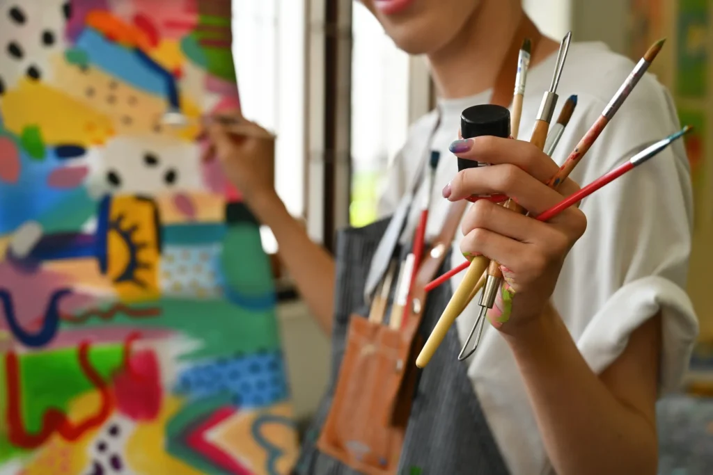 An artist wearing an apron holds several paintbrushes in one hand while painting a colorful abstract artwork on a canvas in a well-lit room.