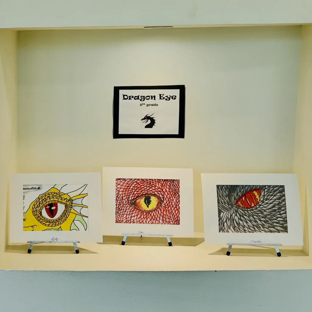 Three dragon eye drawings are displayed on stands below a sign that reads “Dragon Eye 6th Grade,” featuring a dragon silhouette. Each drawing depicts a unique colorful dragon eye in different artistic styles.