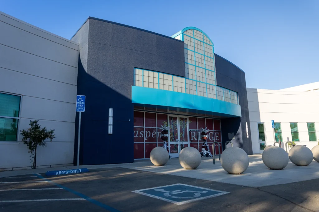 The image shows the entrance of a modern building with a blue and white facade, glass block windows, and large round concrete spheres in front. There are accessible parking spaces and a sign indicating handicapped parking nearby.
