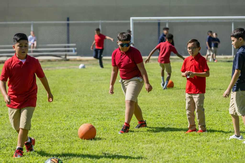 A group of boys wearing red shirts and khaki shorts play soccer with an orange ball on a grassy field with a goalpost and chain-link fence in the background.
