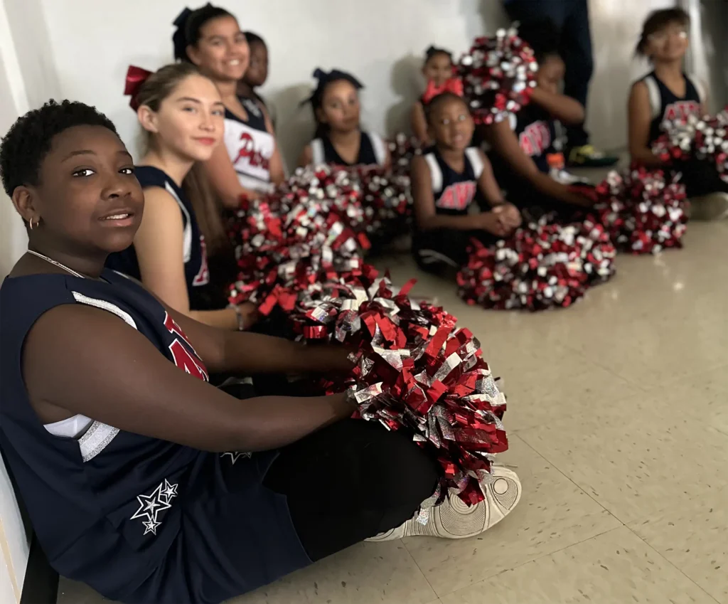 A group of young cheerleaders in matching uniforms sit on the floor indoors, holding red, white, and silver pom-poms. They appear to be waiting, and some are smiling at the camera while others look away.