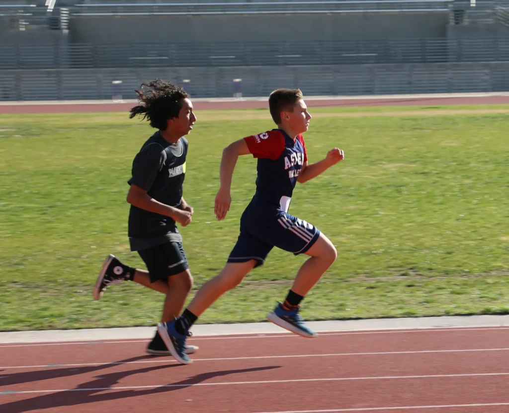 Two boys are sprinting side by side on a running track, both focused and in mid-stride, with a grassy field and stadium stands in the background.