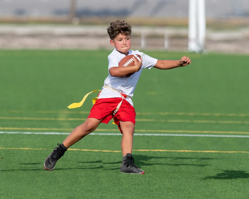 A young boy in a white shirt and red shorts runs on a green field holding a football, wearing black shoes and yellow flag football belts, looking ahead with determination.