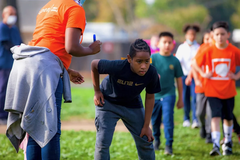 A young boy bends forward, ready to race, while other children and an adult stand behind him on a grassy field. The adult wears an orange shirt and holds a marker. The children are lined up, some wearing orange shirts.