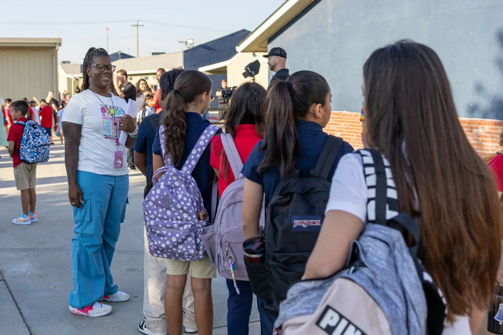 A group of students with backpacks stand in line outdoors at school, while a smiling adult in blue pants and a white T-shirt greets them. Other students and adults are visible in the background.