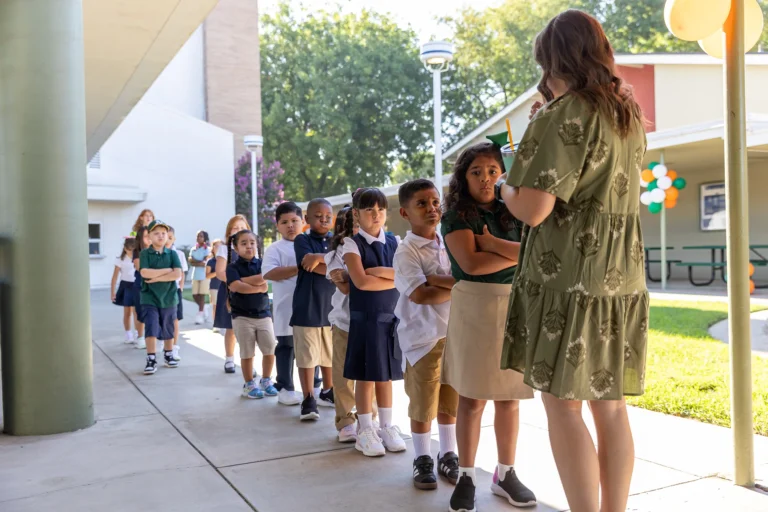 A teacher in a green dress speaks to a group of young students standing in a line outside a school building on a sunny day. The children wear uniforms and carry backpacks.