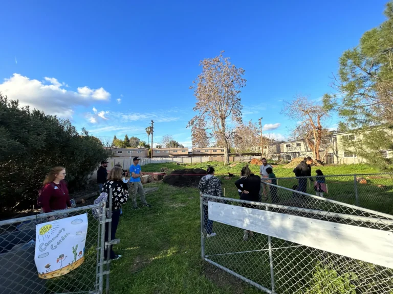 A group of people stands and talks in a grassy, fenced outdoor area with trees and buildings in the background under a bright blue sky. A colorful sign is attached to the fence in the foreground.