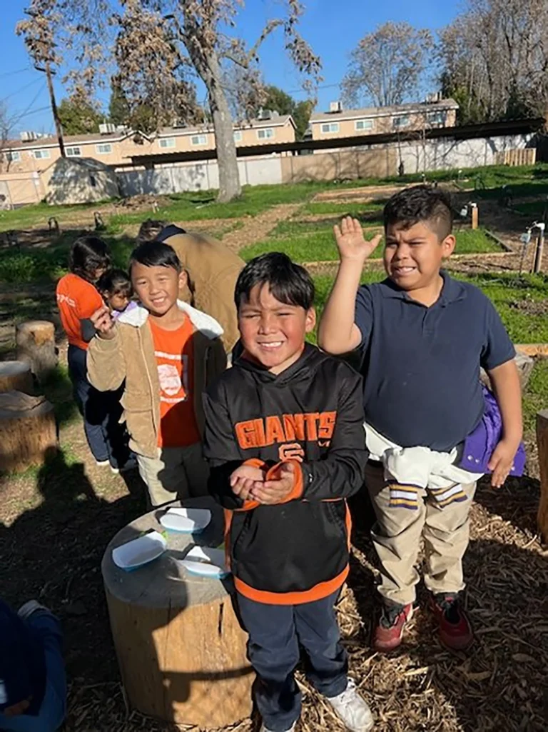 Three smiling boys stand outdoors on a sunny day, with one boy raising his hand. They are surrounded by tree stumps and grass, with other children and buildings visible in the background.