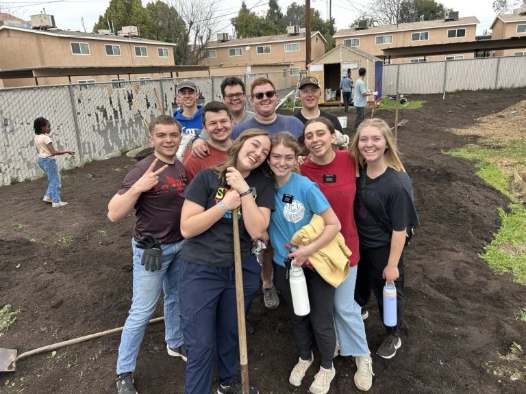 A group of ten smiling young adults pose together outdoors in a garden area, holding tools and water bottles, with residential buildings and a fence in the background. Some are wearing gloves and name tags.