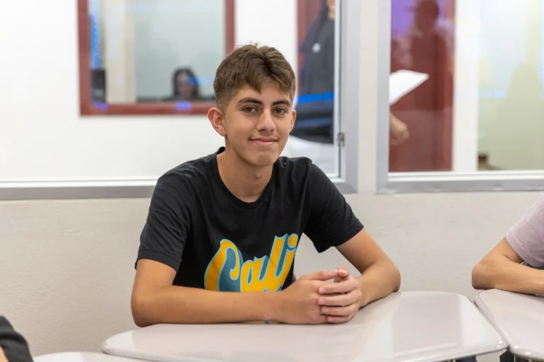 A teenage boy with short brown hair, wearing a black T-shirt that says Cali, sits at a white desk in a classroom, smiling slightly. There are people and windows in the background.