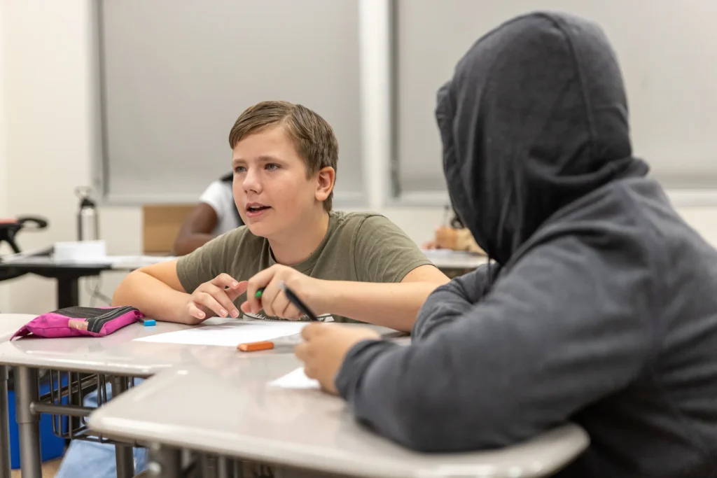 Two students sit at desks in a classroom. One, wearing a green shirt, is speaking, while the other, in a dark hoodie, listens. Papers, a pencil case, and writing materials are on the desks.