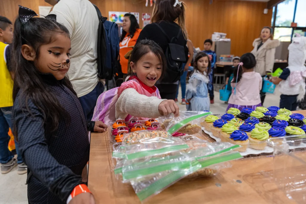 Two young girls in costumes, one with cat whiskers painted on her face, reach for Halloween treats and cupcakes on a table at a party with other children and adults in the background.