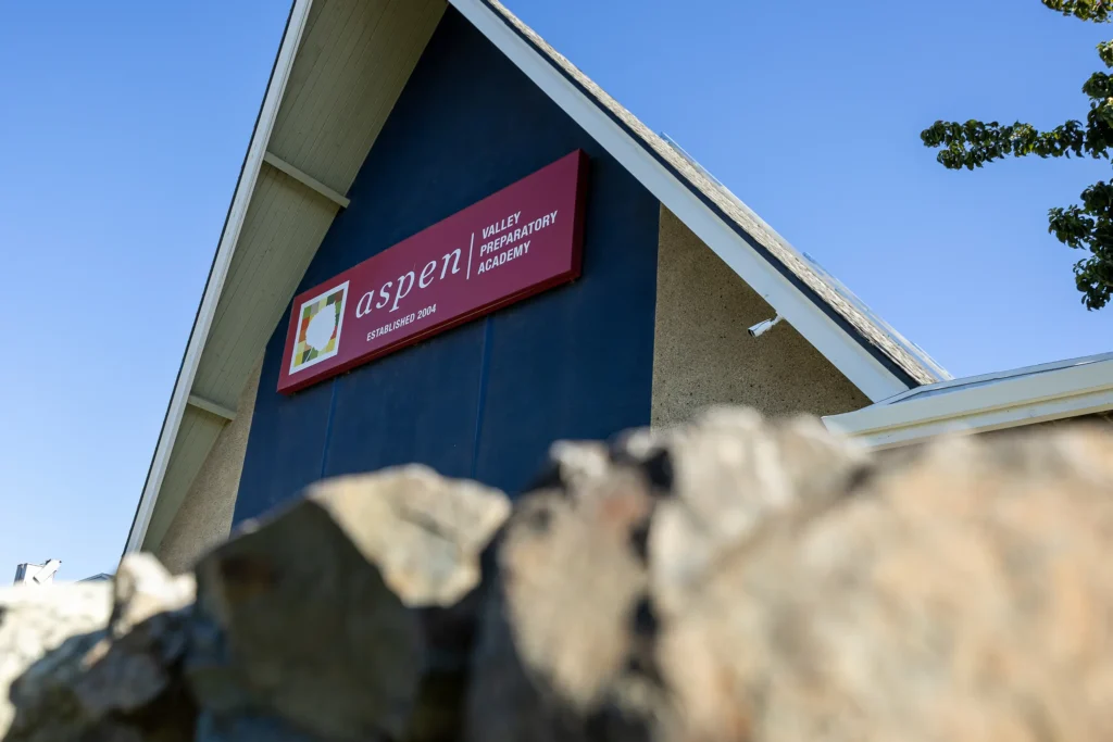 An angled view of the Aspen Valley Preparatory Academy building, partially obscured by rocks in the foreground, with a clear blue sky in the background.
