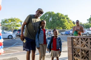 A man in a cap stands with two young girls, both wearing backpacks and school uniforms, outside near a parking lot. Another woman is smiling in the background near a fence and parked cars.