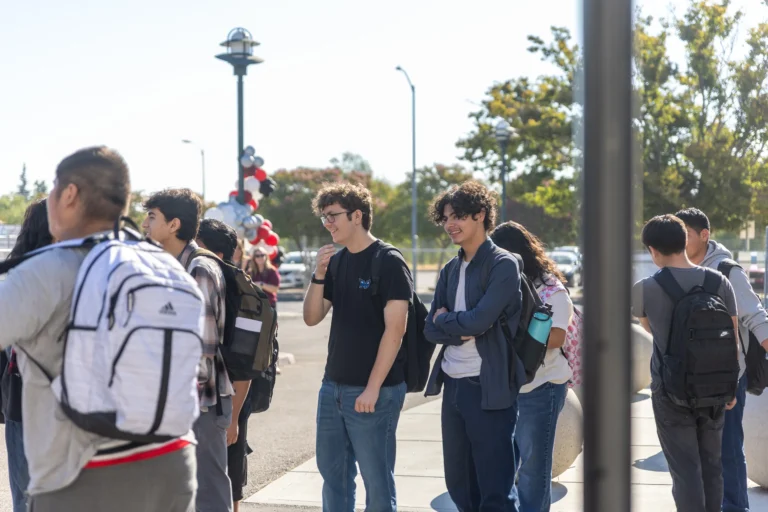A group of students with backpacks stand and talk outside on a sunny day, some smiling and laughing. Trees and balloons can be seen in the background.