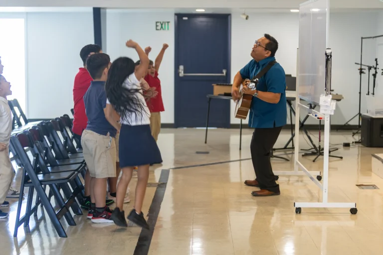 A man plays guitar and sings to a group of children standing and cheering in a bright room with chairs and a whiteboard. The kids appear enthusiastic and engaged with the music.