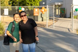 A smiling child with a backpack stands next to a smiling adult in scrubs, holding keys, outside a school entrance decorated with orange, green, and white balloons on a sunny day.