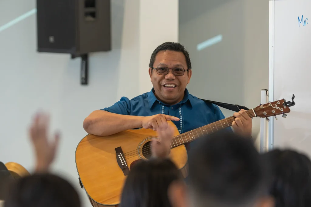 A man wearing glasses and a blue shirt smiles while playing an acoustic guitar in front of a group, with raised hands visible in the foreground.