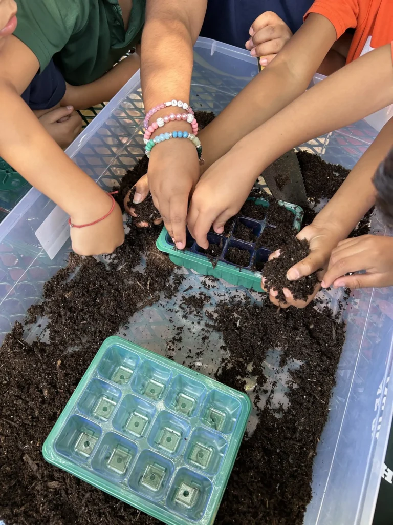 Childrens hands filling small plastic seed trays with soil inside a clear container. Some kids wear colorful beaded bracelets. The activity appears to be a gardening or planting project.