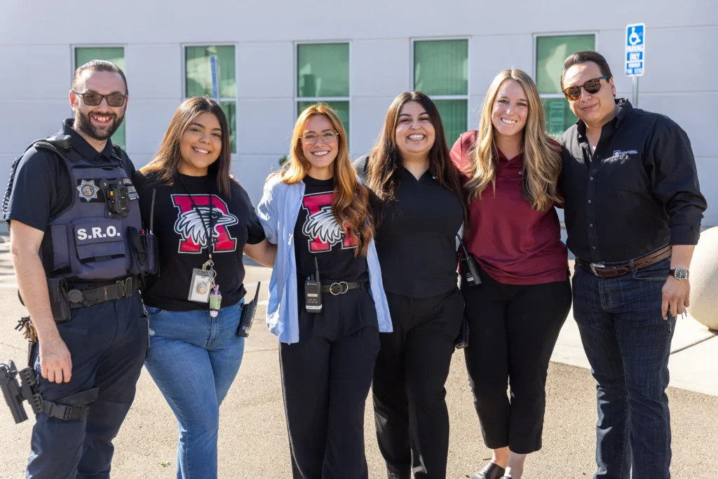 Six adults stand side by side, smiling outdoors. One is dressed as a school resource officer, while others wear casual or business-casual clothing, including two in matching T-shirts with a wildcat logo and the letter “W.”.