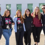 Six adults stand side by side, smiling outdoors. One is dressed as a school resource officer, while others wear casual or business-casual clothing, including two in matching T-shirts with a wildcat logo and the letter “W.”.