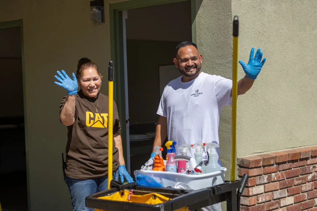Two smiling custodians wearing blue gloves wave while standing next to a cleaning cart with various cleaning supplies, outside a building with an open door.