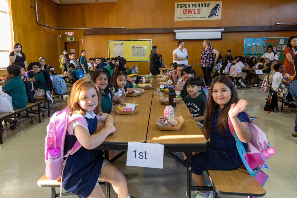 A group of young children sit at cafeteria tables with food trays, smiling and waving. A 1st sign marks their table. Other students and adults are in the background inside a brightly lit school cafeteria.