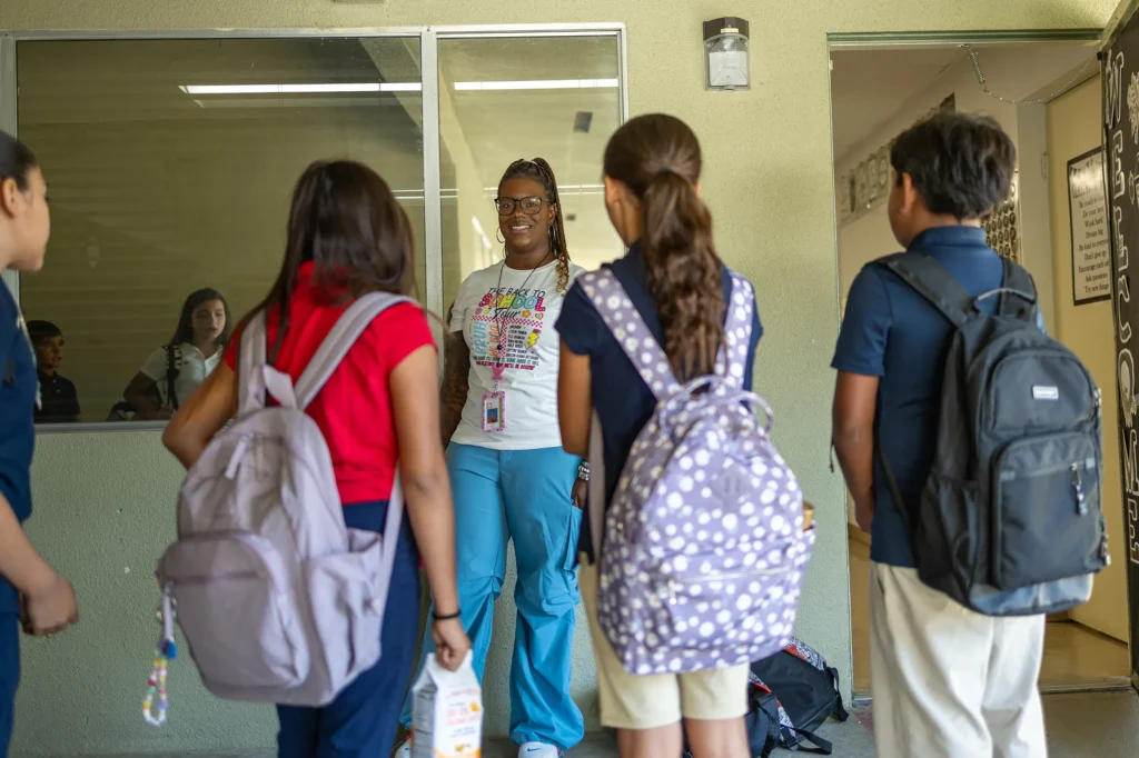 A teacher stands and smiles as she greets a group of four students with backpacks at a school entrance. The students face her, appearing ready for class. A window and open door are visible in the background.