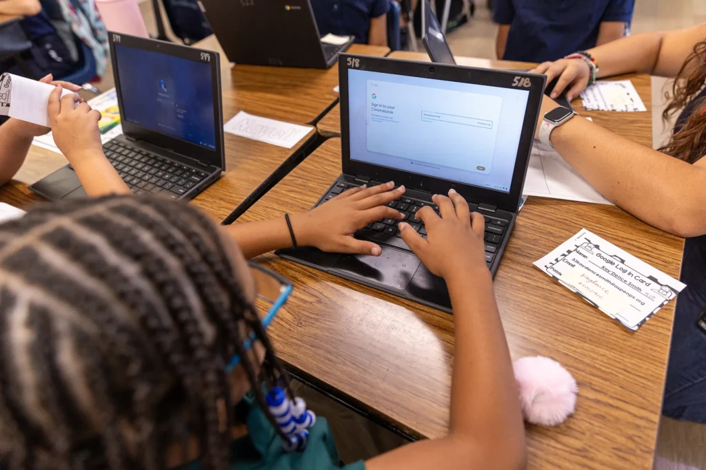 A student with braided hair types on a laptop at a classroom desk, while other students work nearby. Papers, a pink pom-pom keychain, and a teacher’s arm are also visible on the desk.