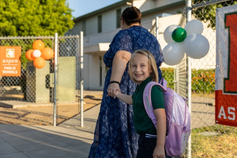 A young girl with a backpack smiles at the camera while holding an adult’s hand, walking into a school entrance decorated with balloons and a sign that reads Aspen Public Schools.