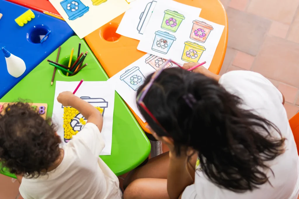 Two children sit at a colorful table, coloring recycling bin drawings with pencils and pom-poms. The table holds art supplies and finished pictures, showing a creative recycling-themed activity.