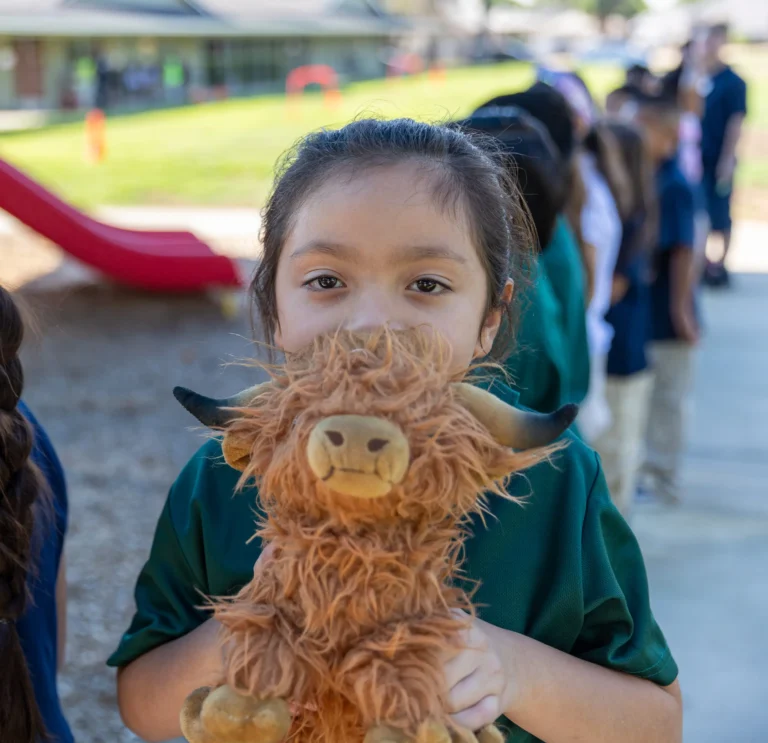 A young girl stands outdoors holding a fluffy brown stuffed animal, with a line of children behind her. She looks directly at the camera, partially hiding her face behind the toy.