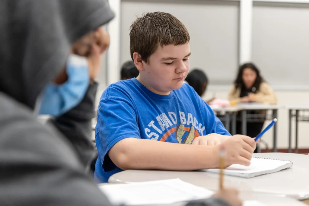 A student in a blue T-shirt writes in a notebook while sitting at a classroom desk, surrounded by other students working in the background.