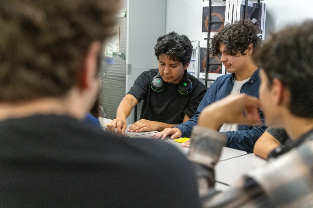 A group of young people sit around a table, focused on a game or activity. One person with headphones around his neck reaches toward the center, while others watch attentively. Shelves and equipment are visible in the background.