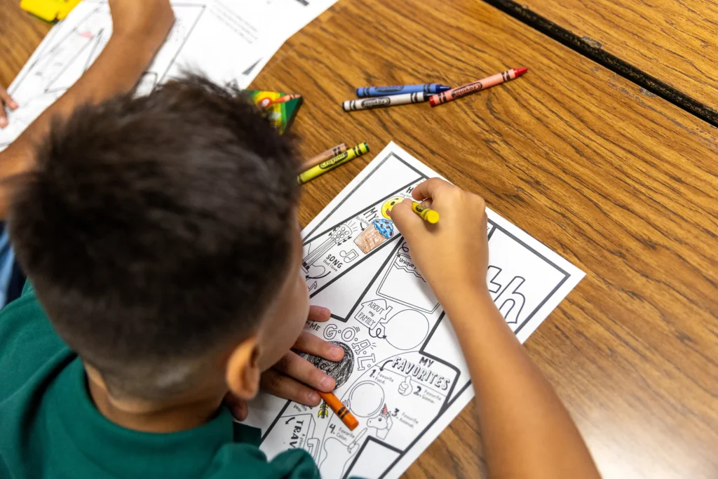 A young child colors a worksheet with crayons at a wooden desk. The worksheet includes various sections and drawings, and several crayons are scattered on the table nearby.