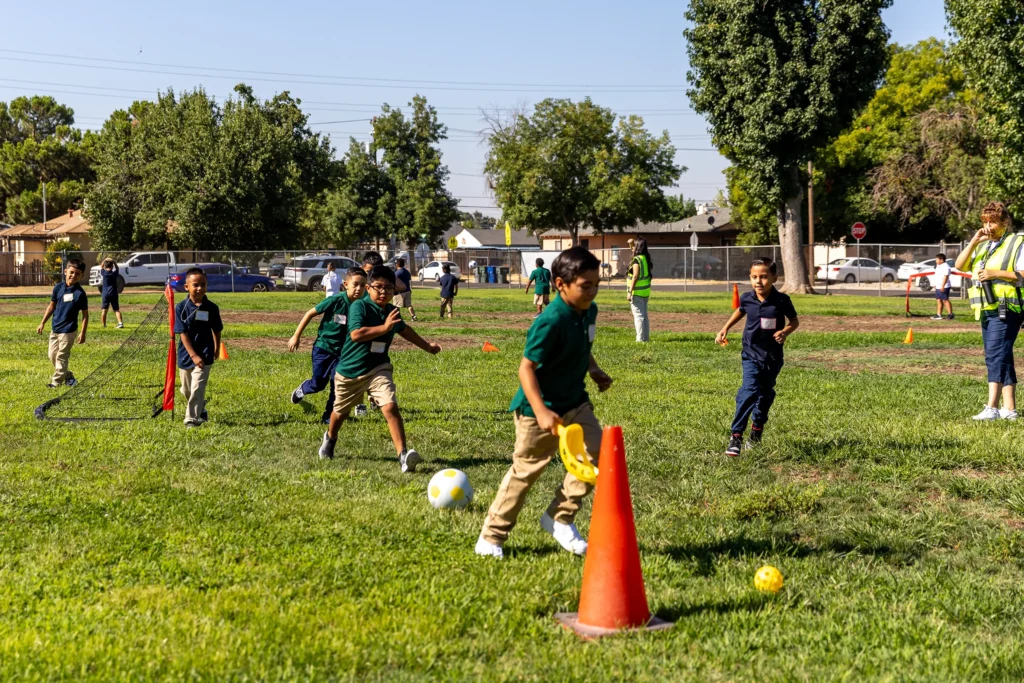 A group of children play soccer on a grassy field, weaving around orange cones, while a few adults wearing safety vests watch nearby. Trees and a fence line the background on a sunny day.